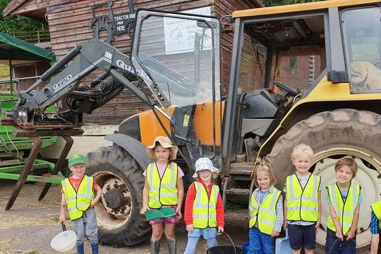 A group of smiling children stands beside a yellow tractor, enjoying a sunny day on the farm