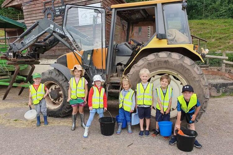 A group of smiling children stands beside a yellow tractor, enjoying a sunny day on the farm